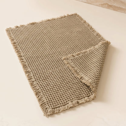 Waffle™ Bath Mat — greige waffle-weave mat with fringed ends on warm cream background