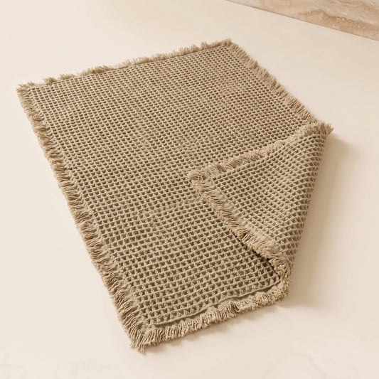 Waffle™ Bath Mat — greige waffle-weave mat with fringed ends on warm cream background