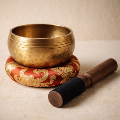 Chant™ Singing Bowl — dimpled copper-alloy bowl resting in its red brocade cushion with the suede-headed mallet, on warm cream plaster